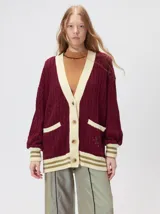 Cardigan de punto trenzado en color negro con detalles en color crema en los bolsillos, puños y ruedo. Presenta un diseño oversize, cierre frontal con botones y bordado decorativo en uno de los bolsillos delanteros.