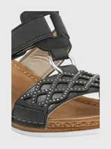 Sandalia Hush Puppies de plataforma baja con diseño de tiras negras. La tira delantera presenta un patrón geométrico calado con pequeñas tachas metálicas, mientras que la tira superior cuenta con un cierre de velcro para un ajuste personalizado. La plantilla es de color marrón y la suela tiene un acabado texturizado que simula madera.