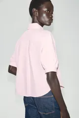 Camisa de popelina color rosado, con cuello redondo amplio, manga corta abullonada y detalle de bordado decorativo con perlas en la botonera frontal oculta.