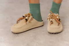 Zueco de cuero gamuzado color tostado, con cordones decorativos adornados con cuentas de madera y piezas envueltas en cuero. Diseño tipo mocasín con costuras visibles en la parte superior y abierto en la parte trasera.