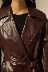 Trinchera de eco-cuero en color bordó, con diseño de doble botonadura, solapas amplias y cinturón ajustable en la cintura. Presenta mangas largas con detalles en los puños y bolsillos laterales.