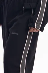 Pantalón deportivo negro de calce relajado con cintura elástica. Presenta una franja lateral gruesa de color gris claro con bordes blancos. Se observa un pequeño bordado en la zona del bolsillo con la palabra "Delosalto".