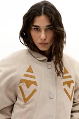 Chaqueta oversized de gamuza de cabra color arena, con apliques tribales en cuero de oveja color mostaza. Presenta hombros exagerados, cuello camisero redondeado, bolsillos frontales y botones a presión forrados. Cuenta con forro capitoneado y un bolsillo interno.