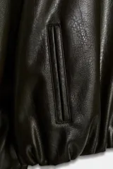 Cazadora negra de efecto piel con cuello solapa, manga larga con elástico en los puños y bajo abullonado con elástico. Cuenta con bolsillos de vivo laterales y cierre frontal con cremallera metálica.