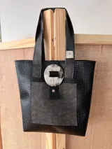Bolso tote negro con textura símil cuero de cocodrilo, bolsillo frontal de cuero liso y cierre con botón metálico.
