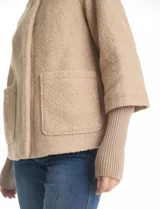 Tapado de tejido boucle color verde oscuro, con silueta evasé y largo a la cadera. Presenta cuello alto envolvente, cierre frontal con botones a presión ocultos y bolsillos frontales de gran tamaño. Cuenta con mangas 3/4 de corte amplio y puños de tejido rib al tono que se extienden para cubrir las muñecas.