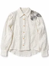 Camisa de lino color crudo, con cuello camisero clásico, mangas largas abullonadas y cierre frontal con botones de madera. Cuenta con un aplique bordado de lentejuelas plateadas en forma de flor sobre el hombro izquierdo.