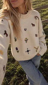Buzo tejido de punto color crudo, con cuello redondo y diseño relajado, adornado con pequeños bordados artesanales multicolor de motivos camperos repartidos por el frente y las mangas.