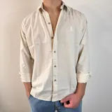 Camisa de manga larga color beige claro, con cuello clásico, cierre frontal con botones y logo bordado en el pecho. Corte regular.