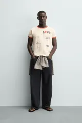 Camiseta de manga corta color crema con cuello redondo y ribetes en color rojo. Presenta un estampado frontal con la palabra "Love" donde la 'o' es un corazón rojo, y varios parches gráficos en tonos grises y negros.