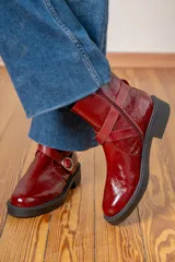 Bota corta de cuero charolado color bordó, con suela plana de goma negra, cierre lateral y tiras decorativas con hebillas metálicas en el tobillo.