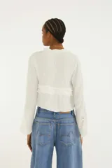 Blusa blanca de manga larga con escote en V, detalles de volados horizontales y cierre frontal con tiras ajustables.