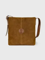 Cartera bandolera de cuero gamuzado color tostado, de diseño cuadrado, con correa larga ajustable y detalle de aplique circular con logo grabado en el centro.