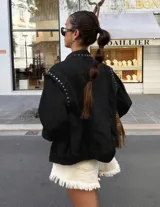 Chaqueta de jean color marrón oscuro, con diseño de hombros caídos y detalle de tachas metálicas en el cuello y la parte superior de las mangas. Presenta cierre frontal con botones metálicos.