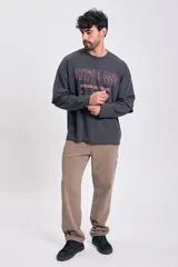 Sudadera de manga larga color gris oscuro con efecto desgastado y dobladillo sin rematar en el bajo. Presenta un diseño de doble manga, donde la manga interior es visible en el puño. Estampado frontal en color rosado con texto temático.