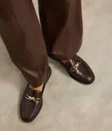 Mocasines de mujer confeccionados en poliuretano color marrón oscuro, con acabado brillante y detalle decorativo metálico dorado en el empeine. Presentan una silueta clásica de punta redondeada y suela plana con pequeño tacón.