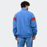 Buzo tipo rompevientos de nylon con medio cierre frontal, cuello alto y puños elásticos. Presenta un diseño en bloques de color azul, blanco y bordó, con el escudo del Club Nacional de Football y el logo de Umbro estampados en el pecho.