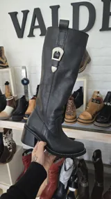 Bota alta de cuero negro con tacón ancho y hebilla metálica decorativa estilo western en el lateral superior de la caña.