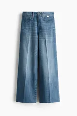 Jean tobillero de denim azul con leve stretch, tiro alto y piernas amplias de corte ancho. Presenta bolsillos de parche delanteros con botón y pliegues marcados.