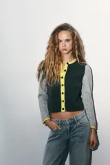 Chaqueta de punto con diseño color block, cuello redondo y manga larga. Presenta cuerpo en color gris oscuro, mangas en gris claro y detalles de ribetes en color amarillo vibrante. Cierre frontal con botones oscuros.