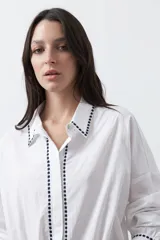 Camisa blanca de manga larga con detalles bordados en contraste a lo largo de la botonadura, el cuello y los puños. Presenta una silueta relajada y un diseño clásico con un toque artesanal.