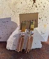 Aros largos colgantes con base rectangular dorada, cuenta con una cuenta facetada y múltiples tiras de strass en tono dorado.