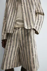 Bermuda de hombre color beige con estampado de rayas verticales azules con efecto desgastado. Corte relaxed fit, con cierre frontal de cremallera y botón, bolsillos delanteros y bolsillos traseros de plastrón.