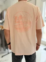 Remera oversize color salmón de manga corta, con cuello redondo y gran estampado circular en la espalda que representa un paisaje de playa con palmeras, olas y el texto circular "May the waves be good where you are".