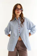 Camisa de corte oversize con estampado de rayas verticales en azul y blanco. Presenta un diseño con recorte diagonal en el frente, aberturas laterales y botones en contraste de color rojo.