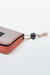 Billetera plegable con cierre de cremallera perimetral. El cuerpo principal es de tela color beige claro, con ribetes y cierre en color salmón/coral. Posee un parche negro con letras blancas en el frente. Incluye un cordón ajustable con tope negro y terminación de cordón en color salmón/coral.