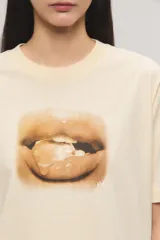 Remera oversize de jersey de algodón color beige claro, con cuello redondo, mangas cortas y estampa frontal de unos labios con un caramelo.