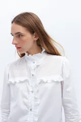 Camisa blanca de algodón con cuello alto con volados y bordado calado en el frente. Tiene mangas largas con puños abotonados y cierre frontal con botones.