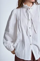 Camisa de color azul con cuello alto decorado con volados y lazo de ajuste. Presenta tablas decorativas en los hombros, cierre frontal con botones de nácar y mangas largas con puños elásticos que generan un efecto abullonado.