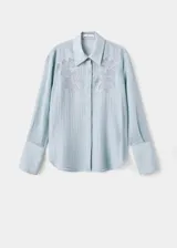 Blusa de tejido ligero color celeste con cuello camisero y cierre frontal de botones ocultos. Presenta mangas largas con puños anchos y detalles florales bordados en la parte superior delantera.