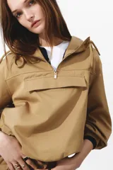 Campera popover color tostado confeccionada en sarga de mezcla de algodón y lyocell. Cuenta con capucha con cordón ajustable, medio cierre frontal y un amplio bolsillo delantero con solapa. Presenta diseño de hombros caídos, canesú frontal, mangas abullonadas con pliegues, puños con cierre ajustable de botón y bajo con cordón interior para fruncir.