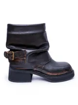 Bota de caña media confeccionada en cuero con efecto desgastado, presenta un diseño holgado en la parte superior y una tira con hebilla metálica ajustable en el tobillo. Cuenta con suela de plataforma y taco cuadrado robusto.