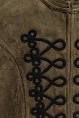 Chaqueta corta de antelina color verde militar, con cuello subido, manga larga y bajo asimétrico. Presenta detalles de alamares negros bordados en contraste en el frente y en los puños.