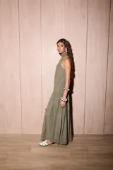 Vestido largo holgado color verde oliva, con escote halter y cuello alto con volado. Presenta un corte con fruncido a la altura de la cadera que genera un maxi volado en la falda.