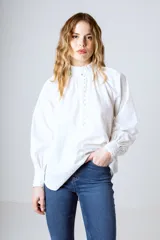 Blusa blanca de algodón con cuello alto con volantes y botonadura frontal parcial. De manga larga con puños abullonados.