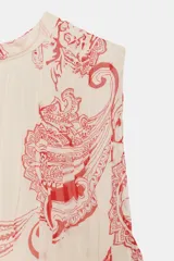 Top sin mangas confeccionado en hilatura de viscosa, con cuello redondo y cierre en la espalda con abertura y botón. Presenta un diseño con estampado paisley a contraste en color rojo sobre fondo beige.