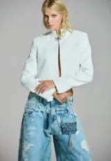 Chaqueta corta de sastrería color blanco, con cuello mao, hombreras marcadas y cierre frontal mediante alamar metálico con diseño de flor y cadena.