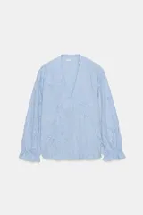Blusa de escote pico y manga larga con puño elástico y volante. Presenta un diseño de rayas verticales finas en azul claro y blanco, con flores en relieve.