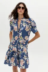 Vestido corto azul de lino con estampado floral en tonos rosados, beige y celestes, cuello mao con escote en V, mangas cortas abullonadas, botones frontales ocultos y volado en el ruedo.