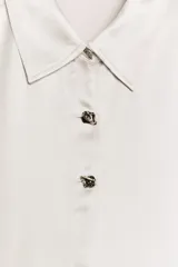 Camisa de cuello solapa y manga larga, confeccionada en tejido satinado con acabado brillante. Presenta cierre frontal con botones y bajo asimétrico.