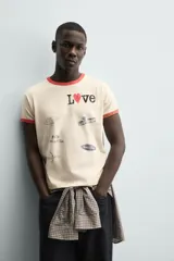Camiseta de manga corta color crema con cuello redondo y ribetes en color rojo. Presenta un estampado frontal con la palabra "Love" donde la 'o' es un corazón rojo, y varios parches gráficos en tonos grises y negros.