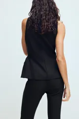 Chaleco entallado de color negro con cuello alto y cierre frontal con cremallera oculta. Presenta un diseño sin mangas con costuras verticales que definen la silueta y un bajo con ligero volumen acampanado.