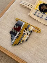 Pin decorativo con estructura de gancho metálico dorado, compuesto por una tira de cuero bordó con costuras en cruz y tres tiras de tela en tonos azul, crema y mostaza. Incluye un parche circular negro con las iniciales BYF bordadas en hilo color ocre.