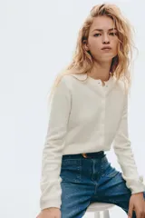 Cárdigan de punto fino, color blanco, de manga larga y corte corto (cropped). Presenta escote redondo y cierre frontal con botones nacarados.