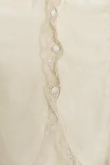 Camisa sin mangas color crema, con cuello redondo y detalle de encaje a tono en el cuello y los bordes de las sisas. Presenta un fruncido horizontal en la parte inferior y cierre con botones forrados en la espalda.