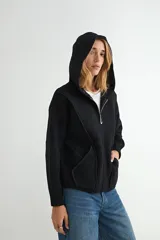 Abrigo negro con capucha, confeccionado en tejido de textura suave. Presenta cierre frontal con cremallera, bolsillos laterales integrados con costuras decorativas y puños ajustados.